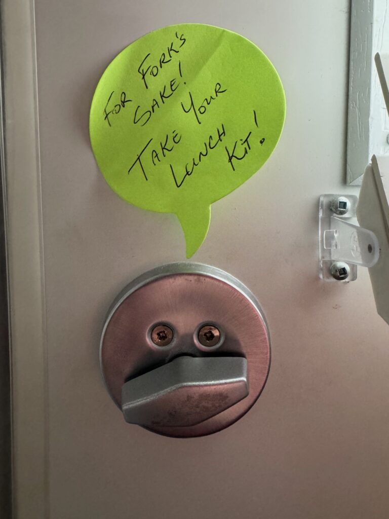 Bright green word bubble shaped sticky note above inside lock on white door. The note reads "For fork's sake, take your lunch kit!"