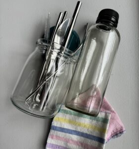 Two clear glass jars, one filled with eating utensils and reusable straws, lie on their sides. At the bottom of the image is one striped cloth napkin and one pink multipurpose cloth.