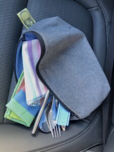 A zero-waste lunch kit consisting of a grey wool zippered pouch, striped cloth napkins, and various eating utensils. The kit is sitting on a car seat and is viewed from above. The SD part of the Something Different Upcycled logo in-seam tag is visible in the top left of the image.